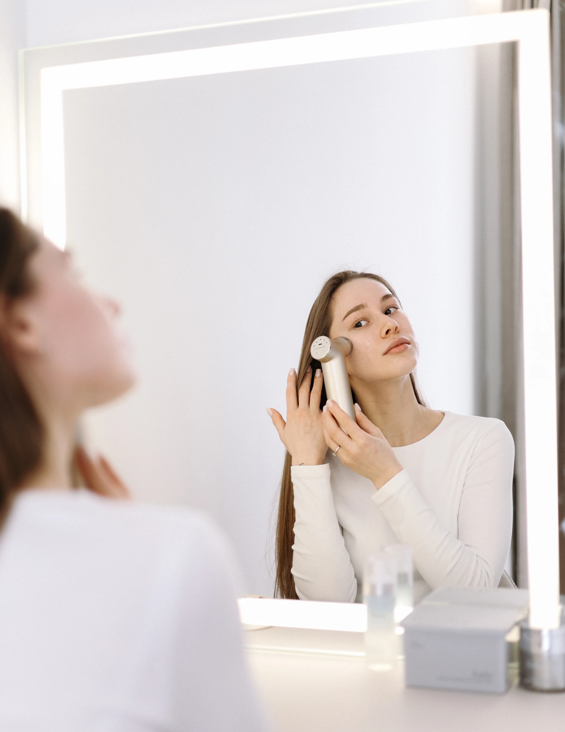 A woman looks in a lighted mirror while using a handheld beauty device on her face.