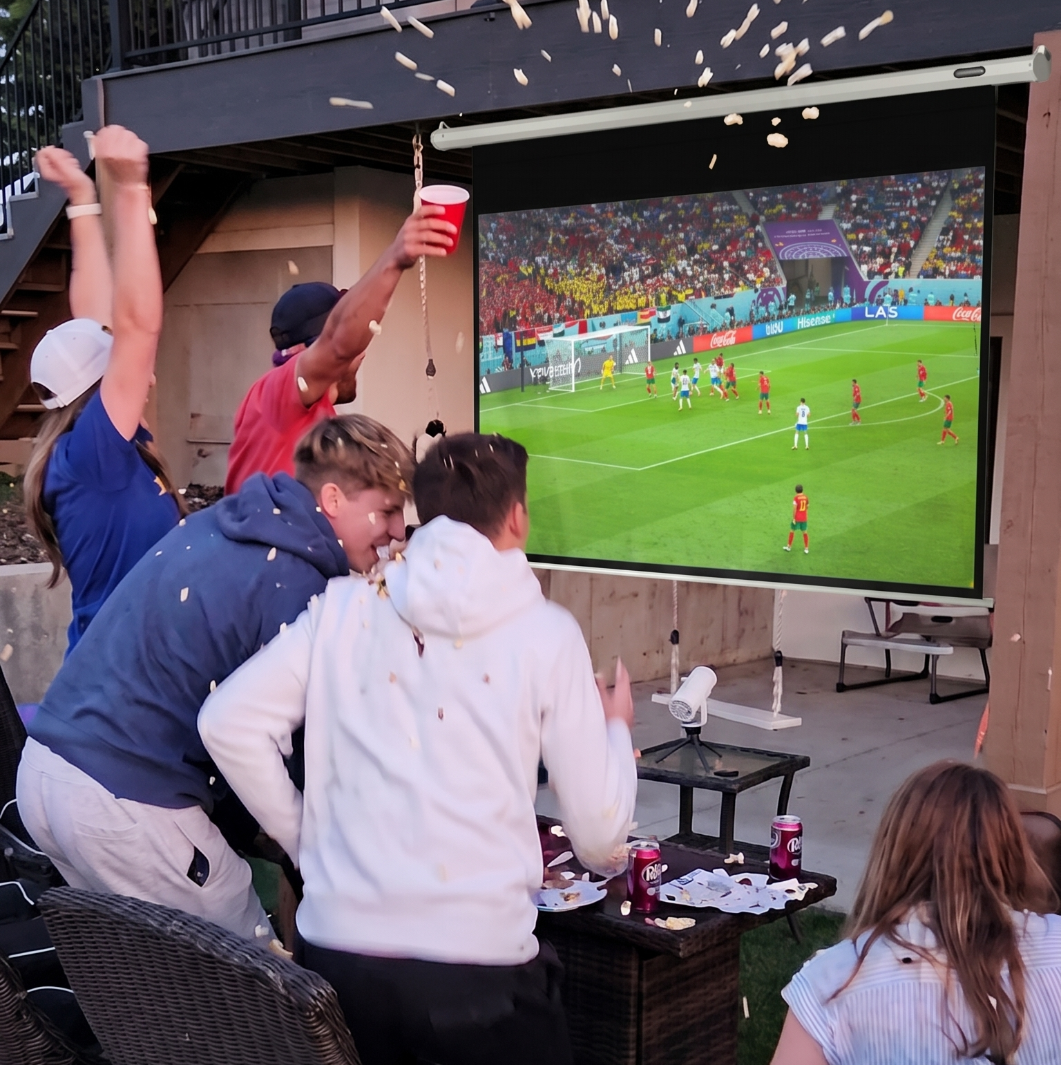 A group of people celebrates by throwing popcorn while watching a soccer game on an outdoor projector screen.