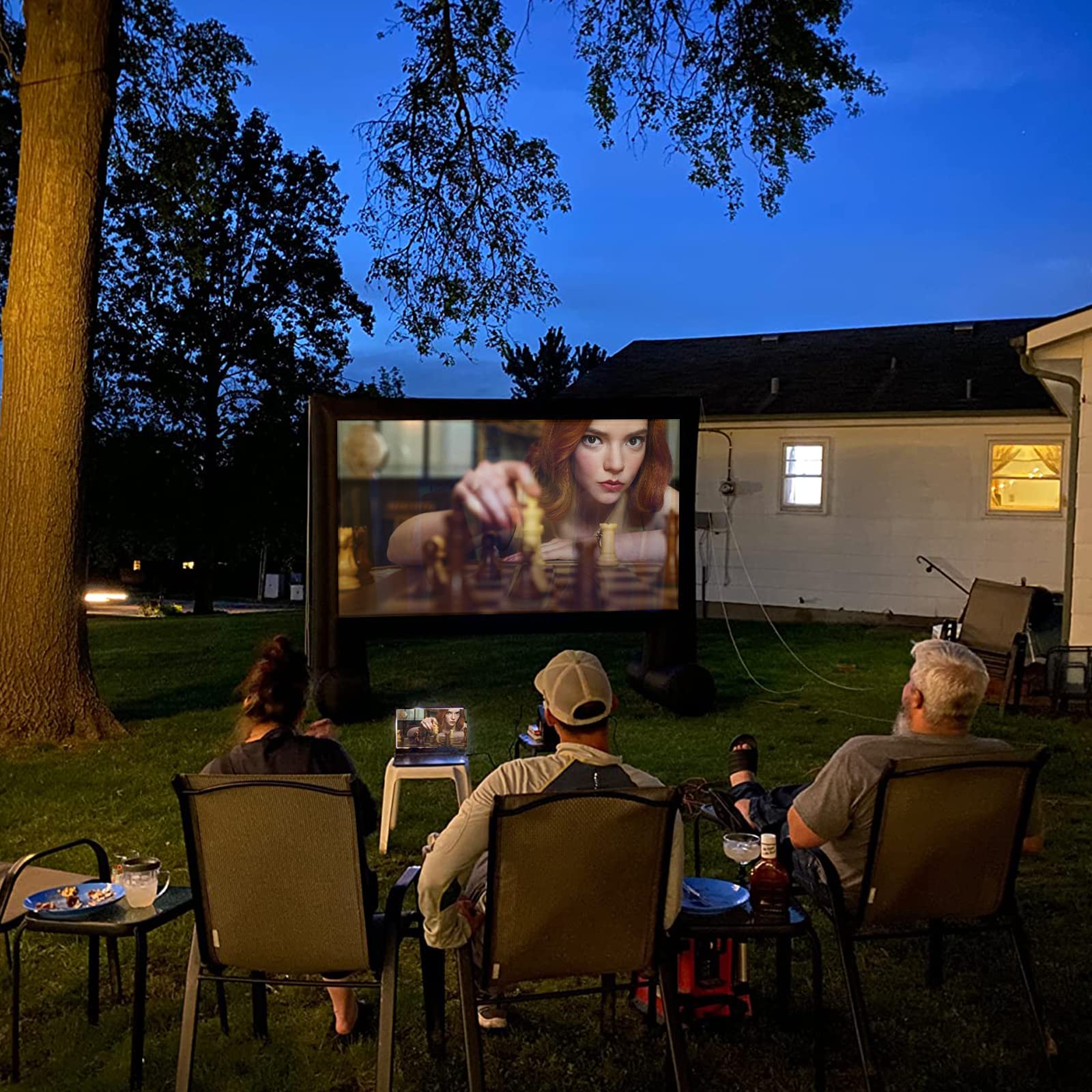 Three people watching a movie on an outdoor screen in a backyard at dusk.
