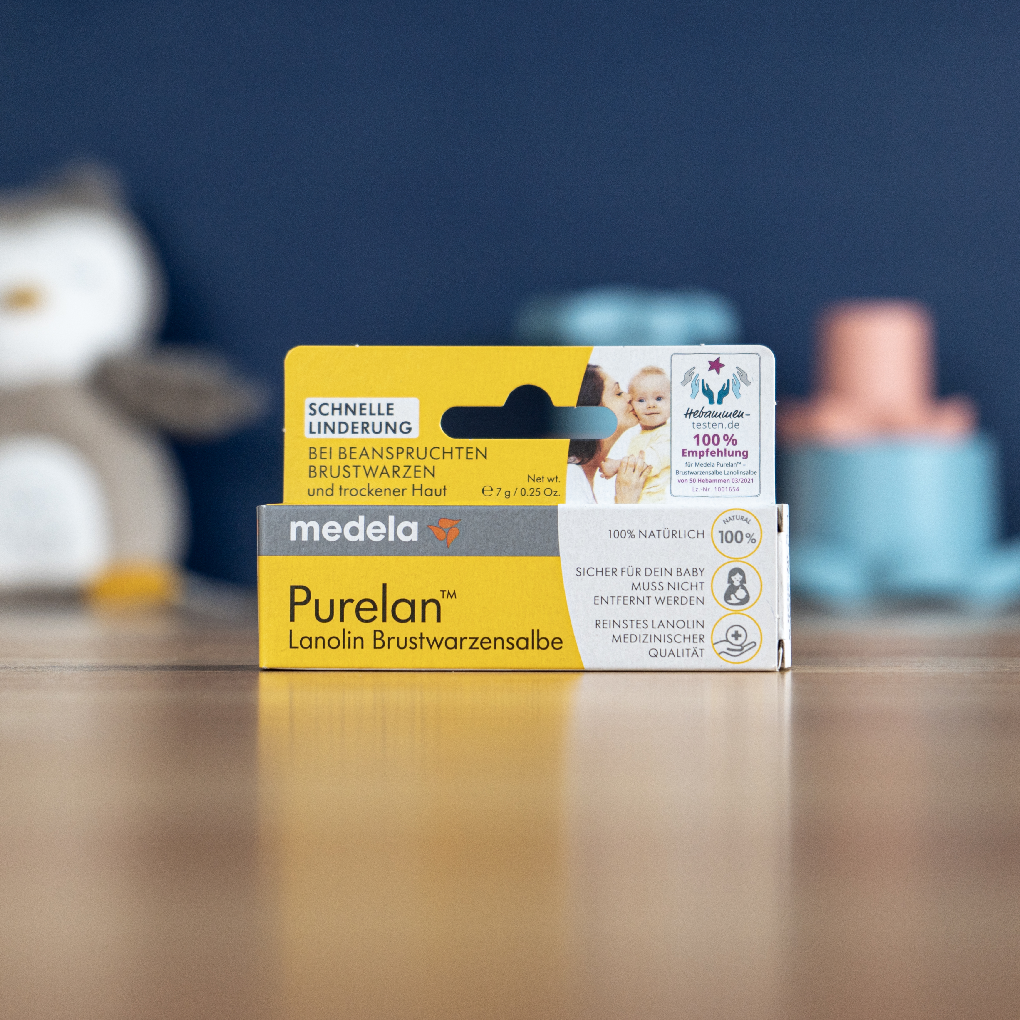 A small yellow and white box of Medela Purelan ointment on a wooden surface with toys blurred behind.
