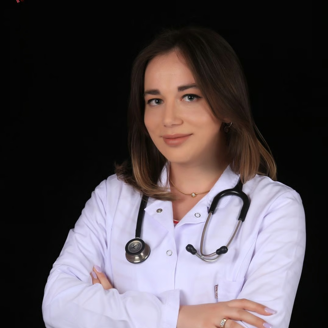 A female doctor in a white coat with a stethoscope around her neck, posing with her arms crossed.