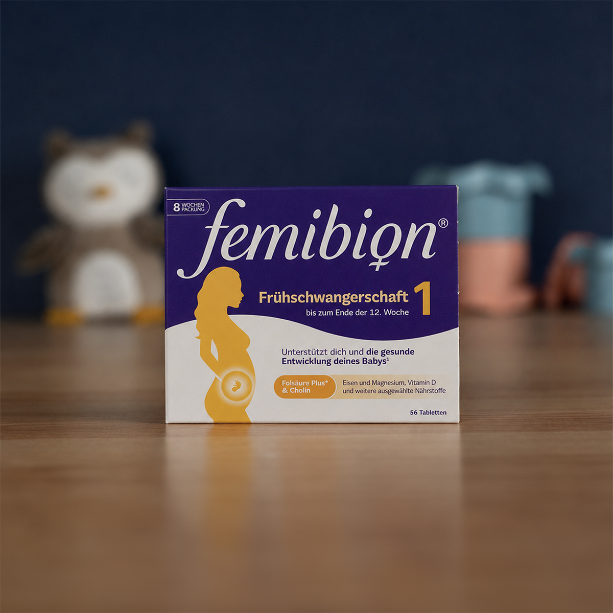 A box of Femibion 1 Frühschwangerschaft prenatal vitamins on a wooden table, with toys blurred in the background.