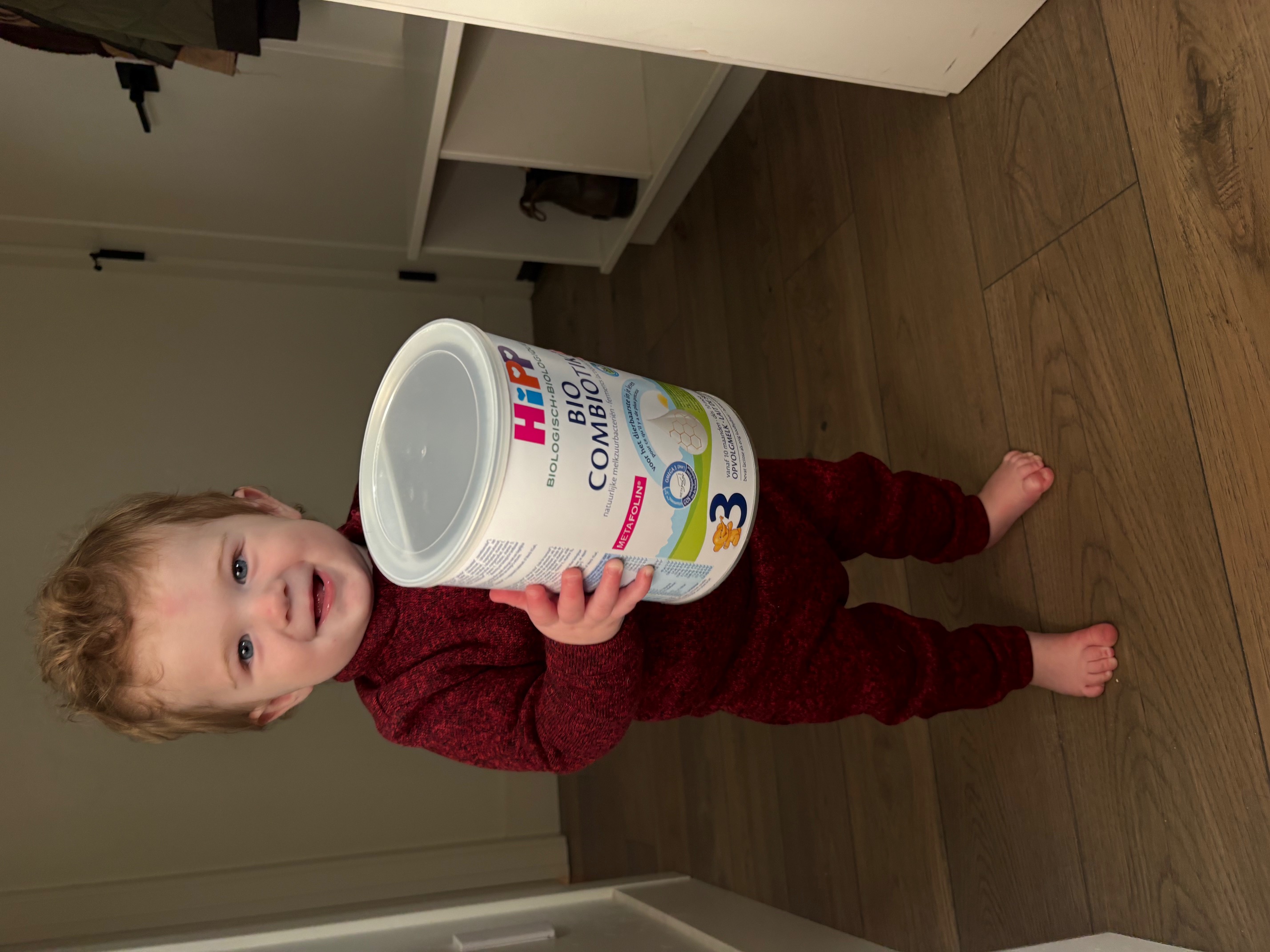 A smiling toddler in a red knit outfit stands barefoot, holding a large can of HiPP baby formula.