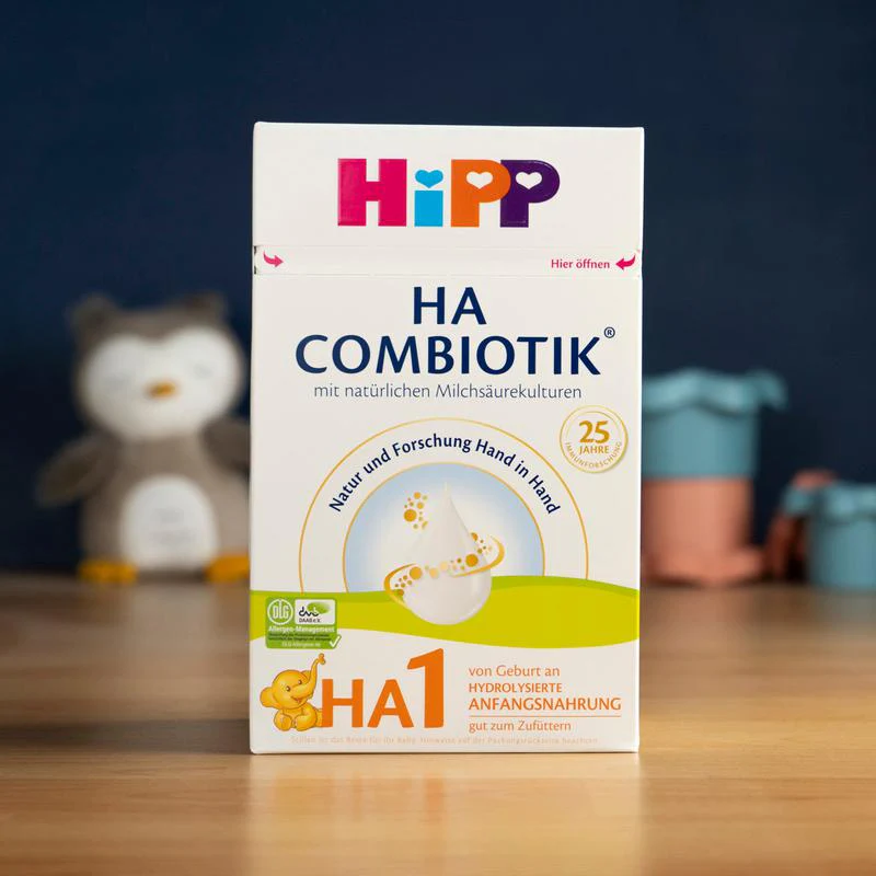 A box of HiPP HA Combiotik baby formula sits on a wooden table, with a stuffed animal blurred behind it.