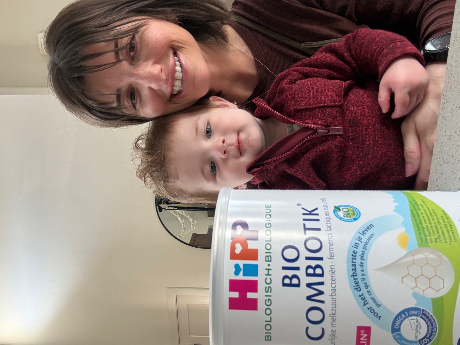 A smiling woman holds a small child next to a can of HiPP Bio Combiotik formula.