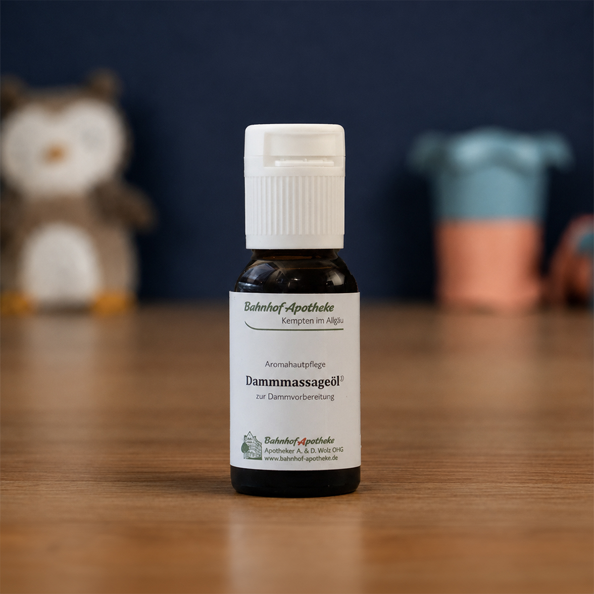 A small brown bottle of Bahnhof-Apotheke Dammmassageöl sits on a wooden table with blurred toys in the background.