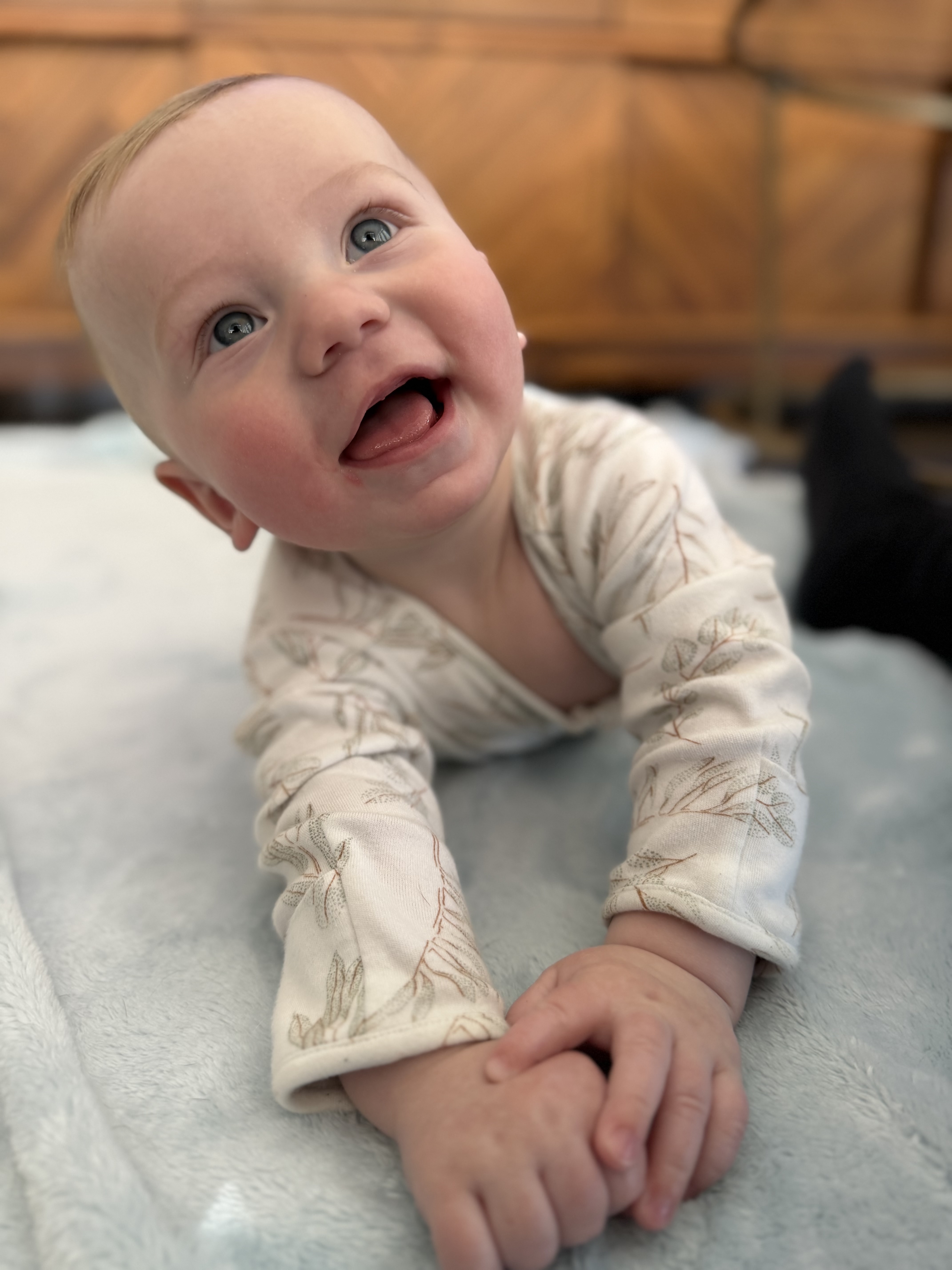 A smiling baby with blue eyes is lying on its tummy on a light blue blanket, looking up.