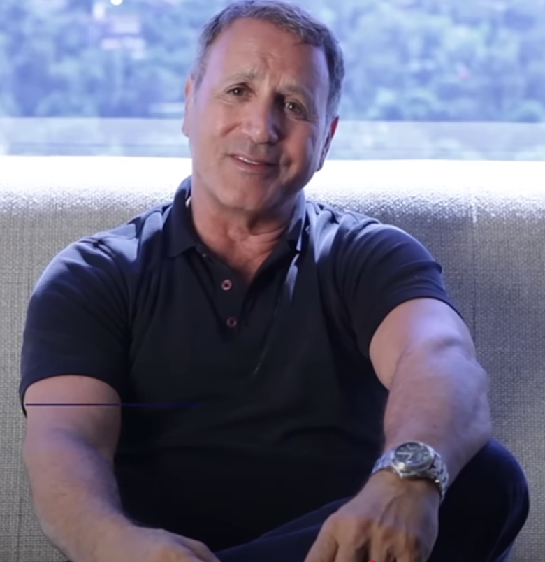 Man sitting on a couch, wearing a black shirt and watch, with a blurred background.