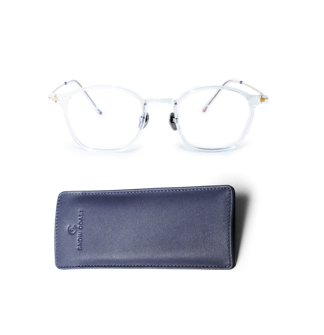 A pair of clear-framed eyeglasses with metal arms rests above a blue leather slipcase on a white background.