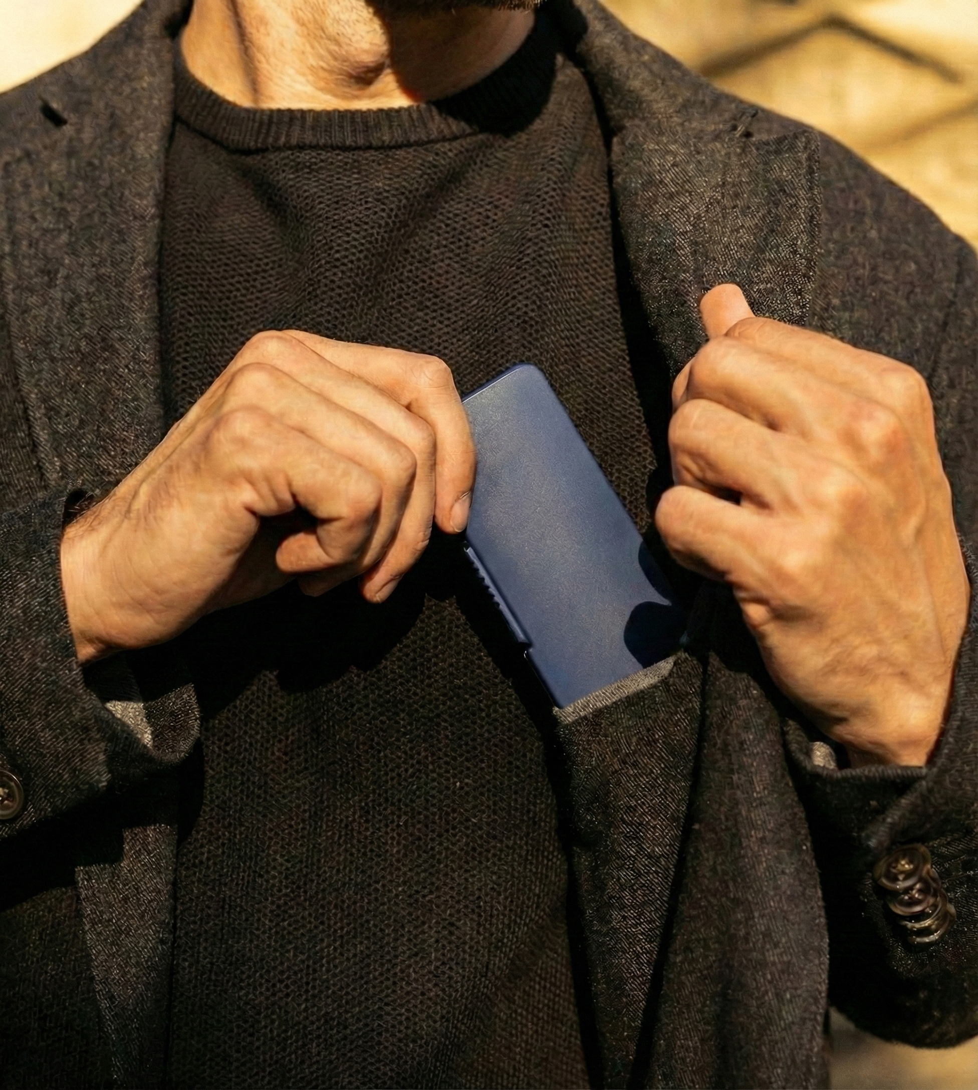 Close-up of a man in a textured brown blazer sliding a slim navy blue glasses case into his interior breast pocket.