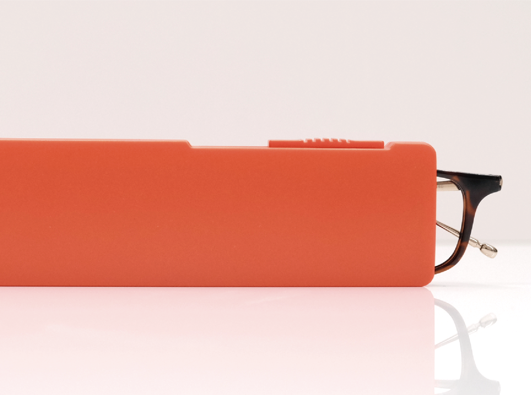 A pair of tortoiseshell glasses sits partially inside a flat, orange case on a reflective white surface.