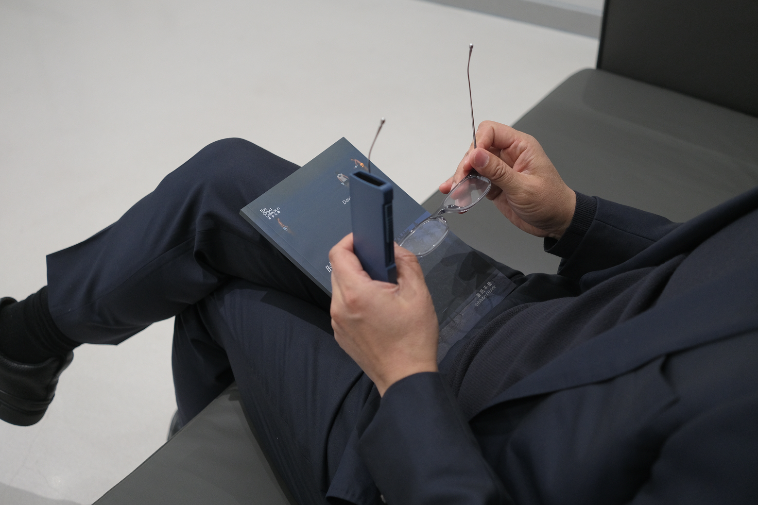 Top-down view of a person holding clear rectangular reading glasses and a slim blue protective case while reading a book.
