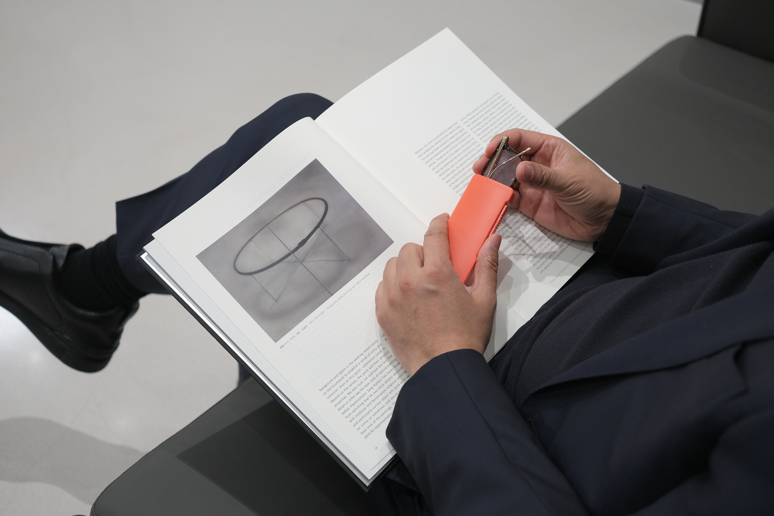 Person holding a vibrant orange ultra-slim glasses case while reading an art book, showing the compact lifestyle design.