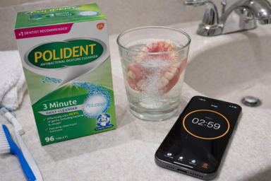 A box of Polident next to dentures soaking in a glass, with a smartphone timer counting down.