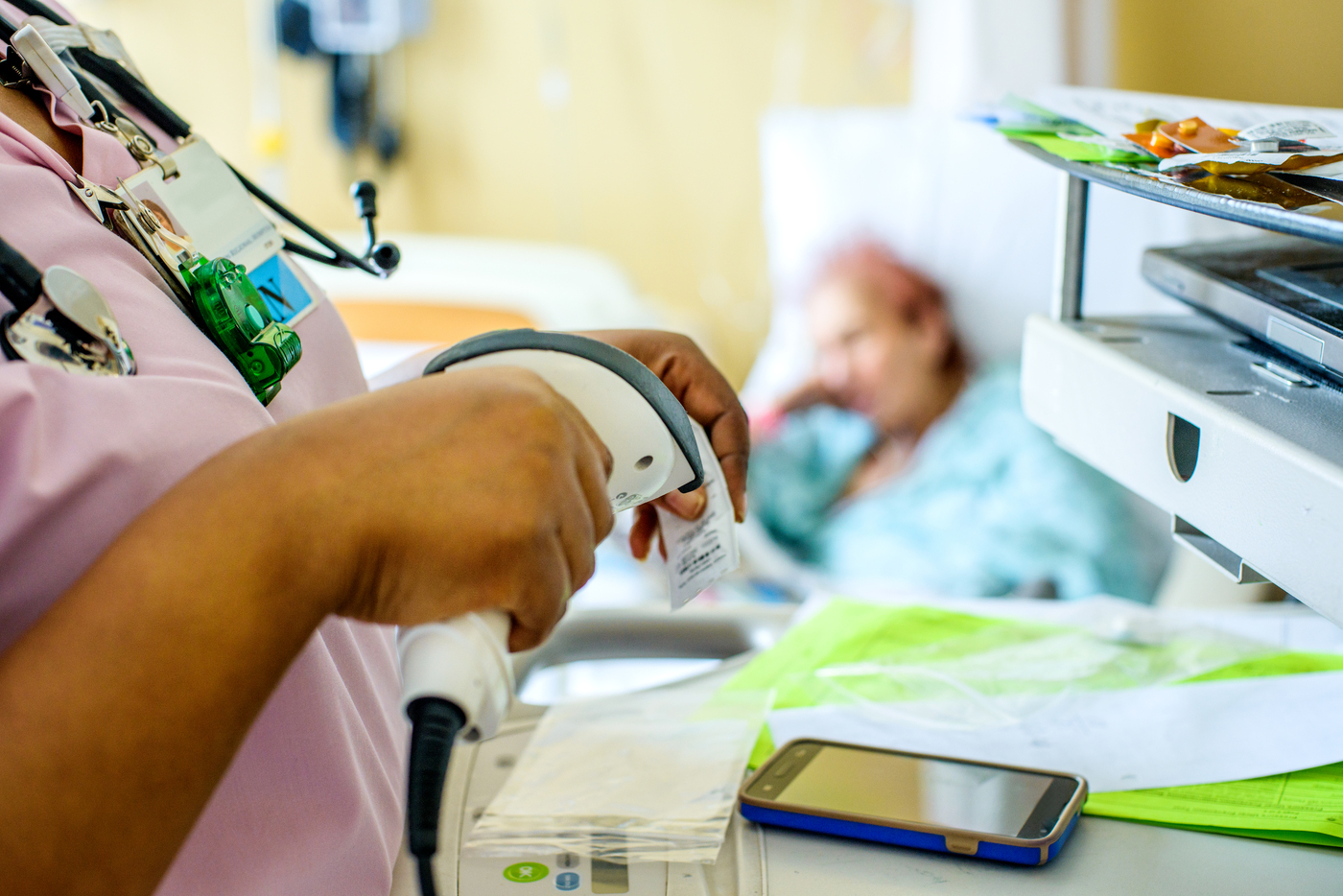 A nurse scans a barcode in a hospital room with a patient lying in a bed in the background.