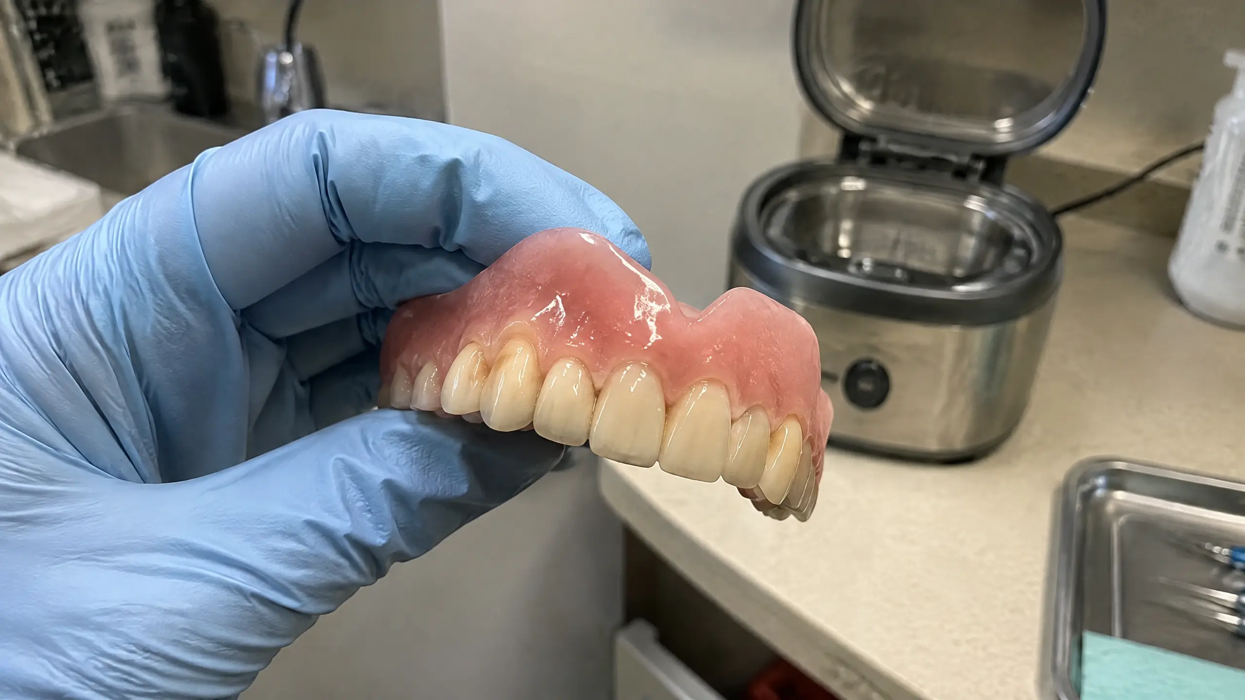 A person wearing a blue medical glove holds a set of upper dentures in a dental office.
