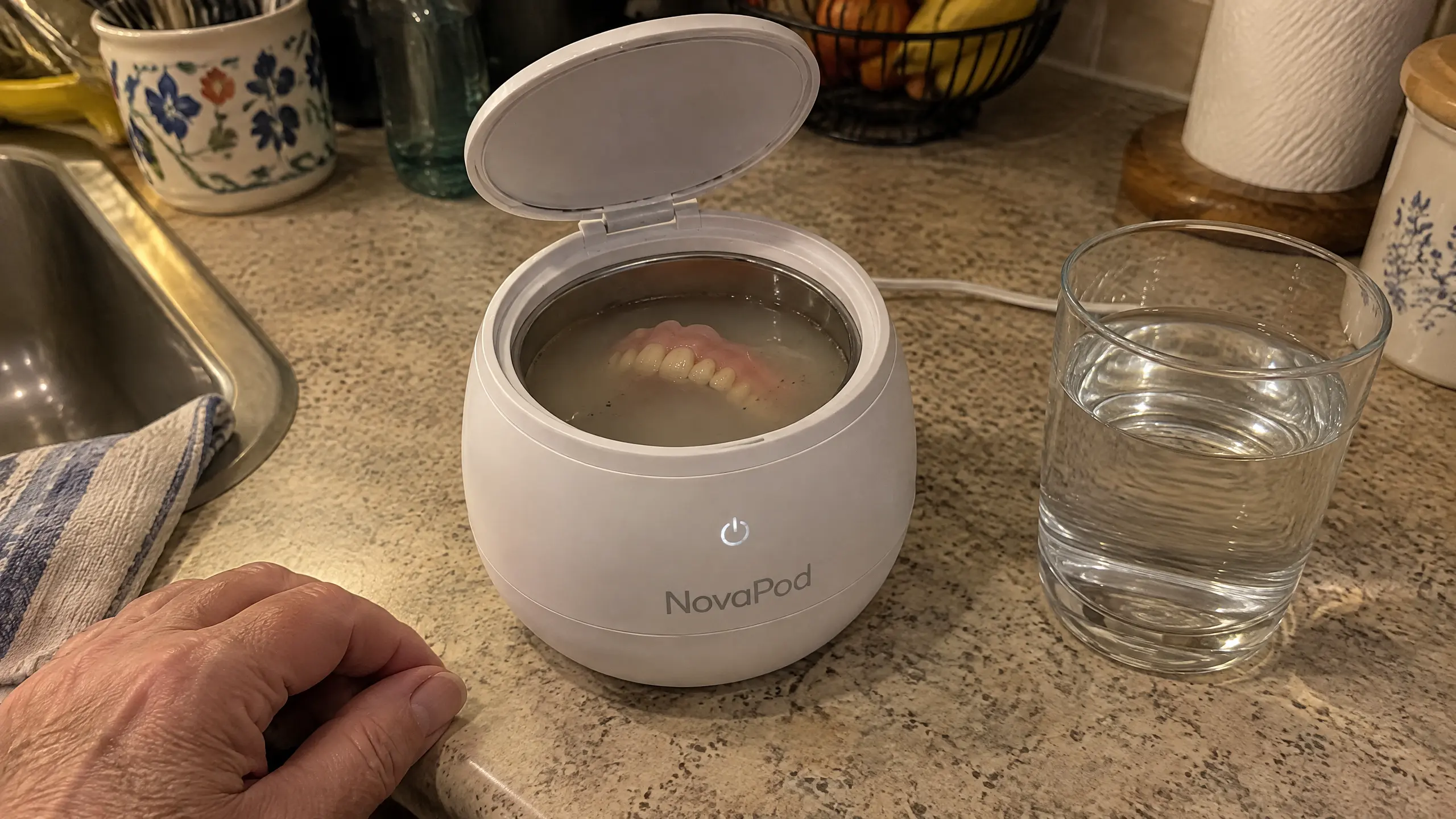 A set of dentures soaking in a white NovaPod ultrasonic cleaner on a kitchen counter.