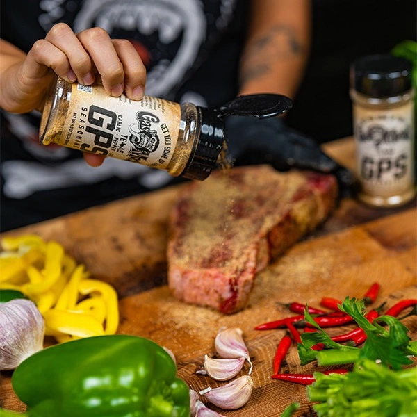 A person sprinkles seasoning from a shaker onto a raw steak surrounded by fresh peppers and garlic.