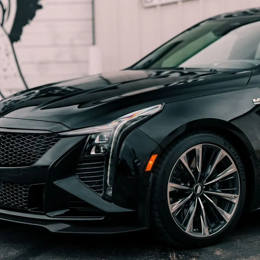 A close-up of the front quarter of a sleek black car, showing its headlight, grille, and wheel.