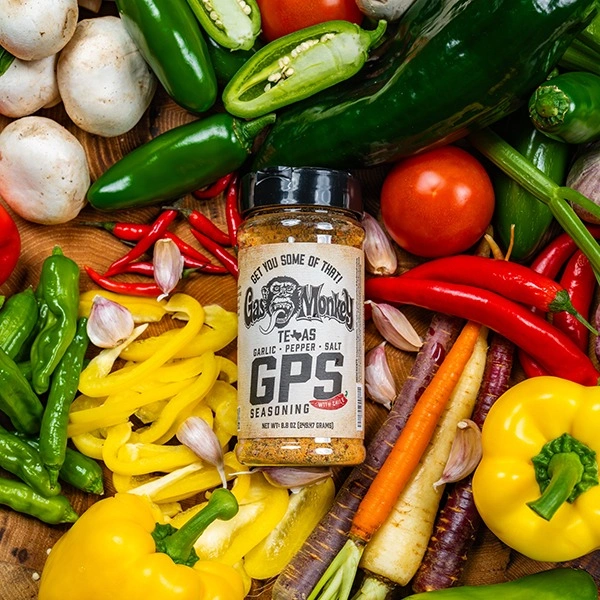 A bottle of Gas Monkey GPS seasoning surrounded by fresh peppers, garlic, mushrooms, and carrots on a wooden surface.