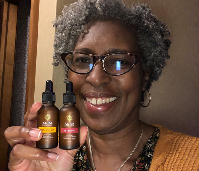 A smiling woman with gray hair and glasses holds up two small bottles of Aura Naturals essential oils.