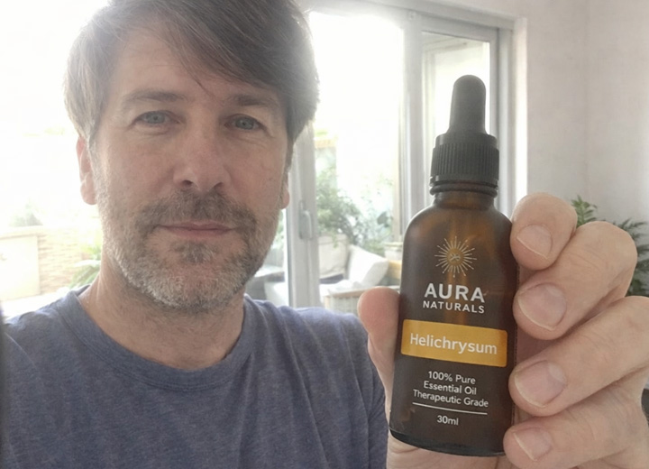 A man holds up a bottle of Aura Naturals Helichrysum essential oil while taking a selfie.