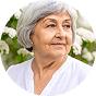 Elderly woman with gray hair, wearing a white shirt, standing in front of white flowers.