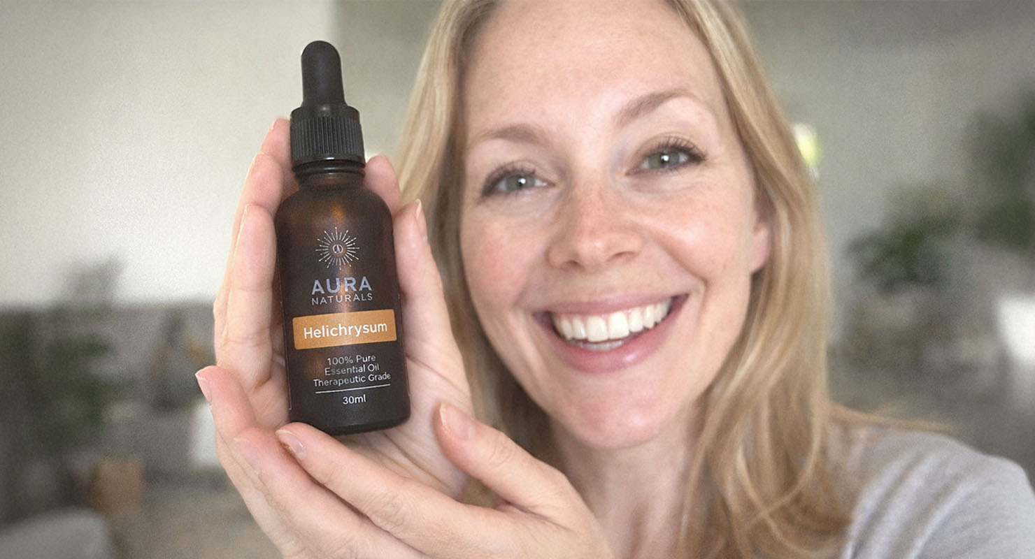 A smiling woman holds a bottle of Aura Naturals Helichrysum essential oil close to the camera.