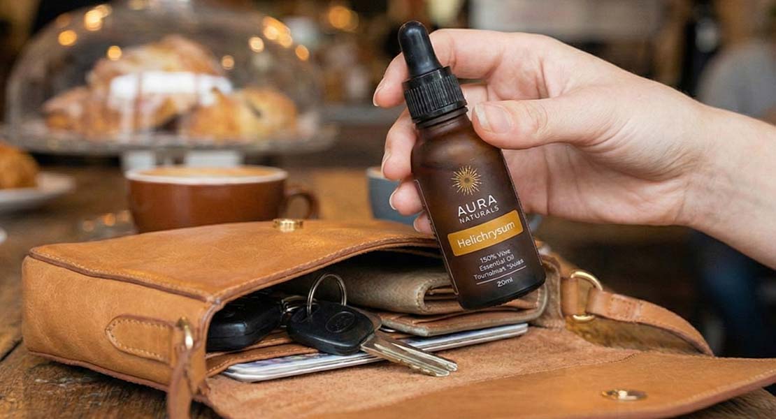 A hand holds a small brown bottle of Aura Naturals Helichrysum oil over an open brown leather purse.