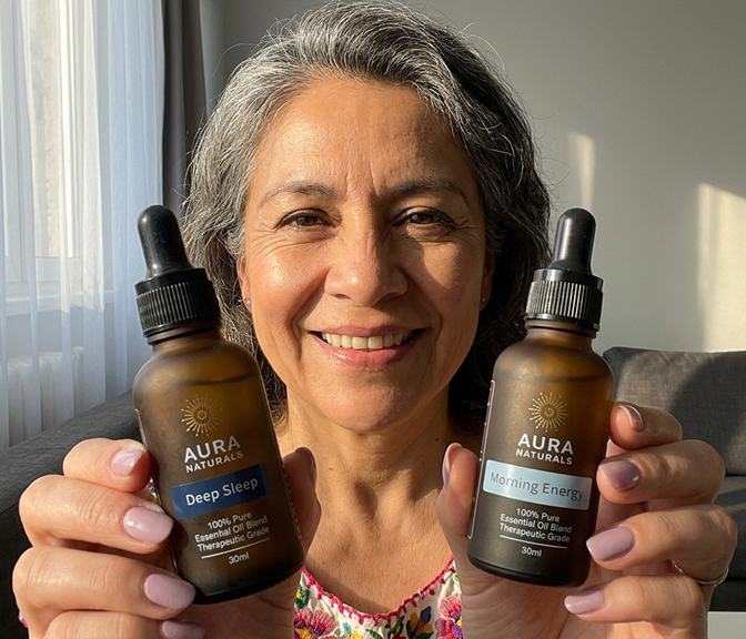 A smiling woman with gray hair holds up two Aura Naturals essential oil bottles.
