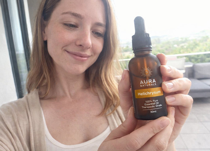 A smiling woman holds up a bottle of Aura Naturals Helichrysum essential oil.