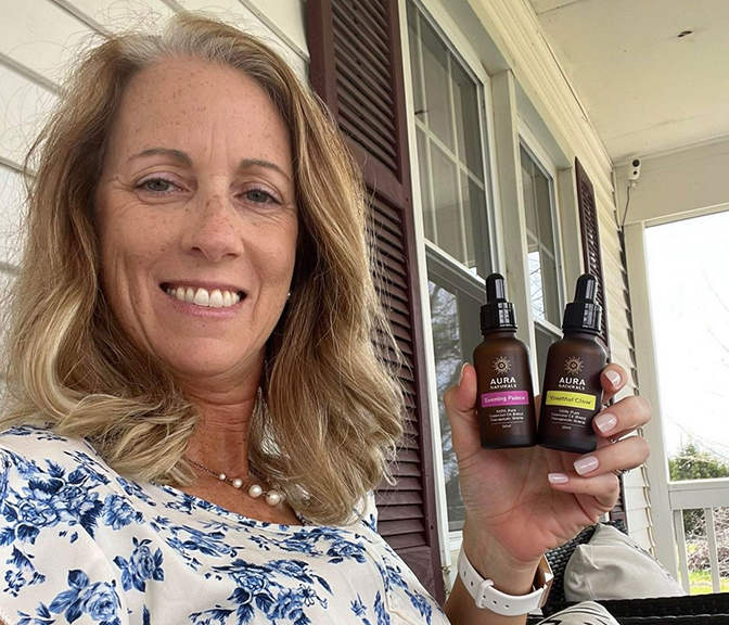 A smiling woman with blonde hair holds up two small brown dropper bottles while sitting on a porch.