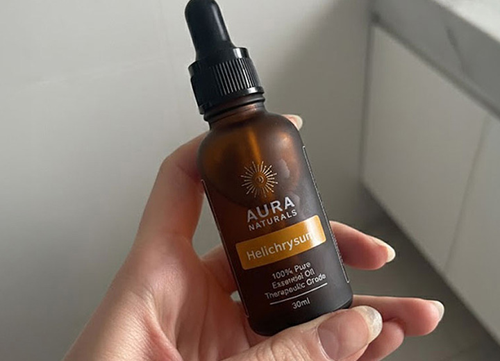 A hand holds a brown dropper bottle of Aura Naturals Helichrysum essential oil against a white background.