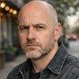 A headshot of a bald man with a short beard and a serious expression, wearing a dark jacket.