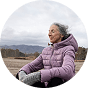 Elderly woman in a purple jacket sitting outdoors with mountains in the background.