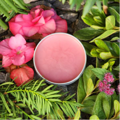 An open round tin of pink balm, surrounded by pink flowers and green foliage on dark rocks.