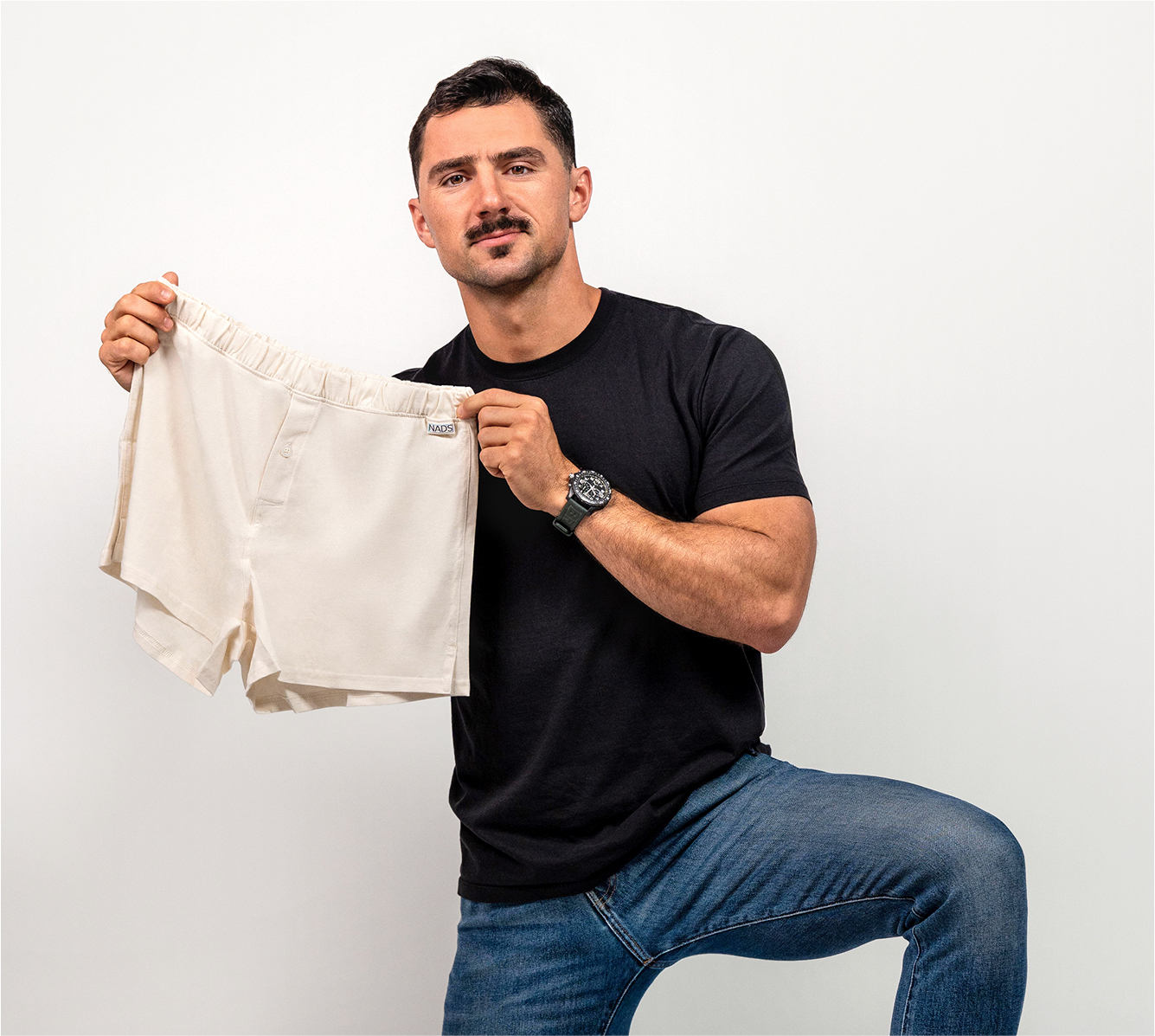 Man in black shirt holding beige shorts against a plain background.