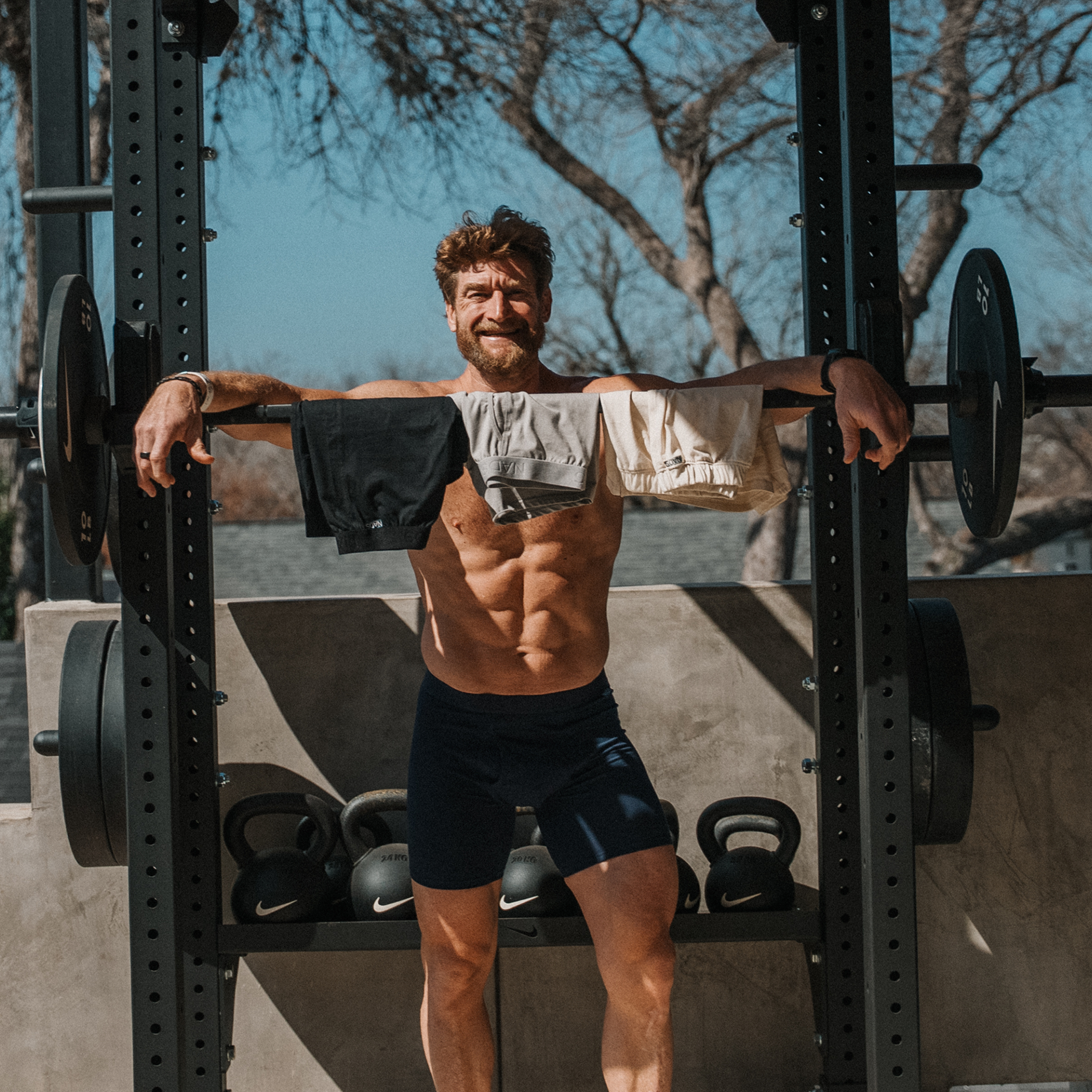 A muscular, shirtless man holds a barbell with three pairs of shorts draped over it in an outdoor gym.