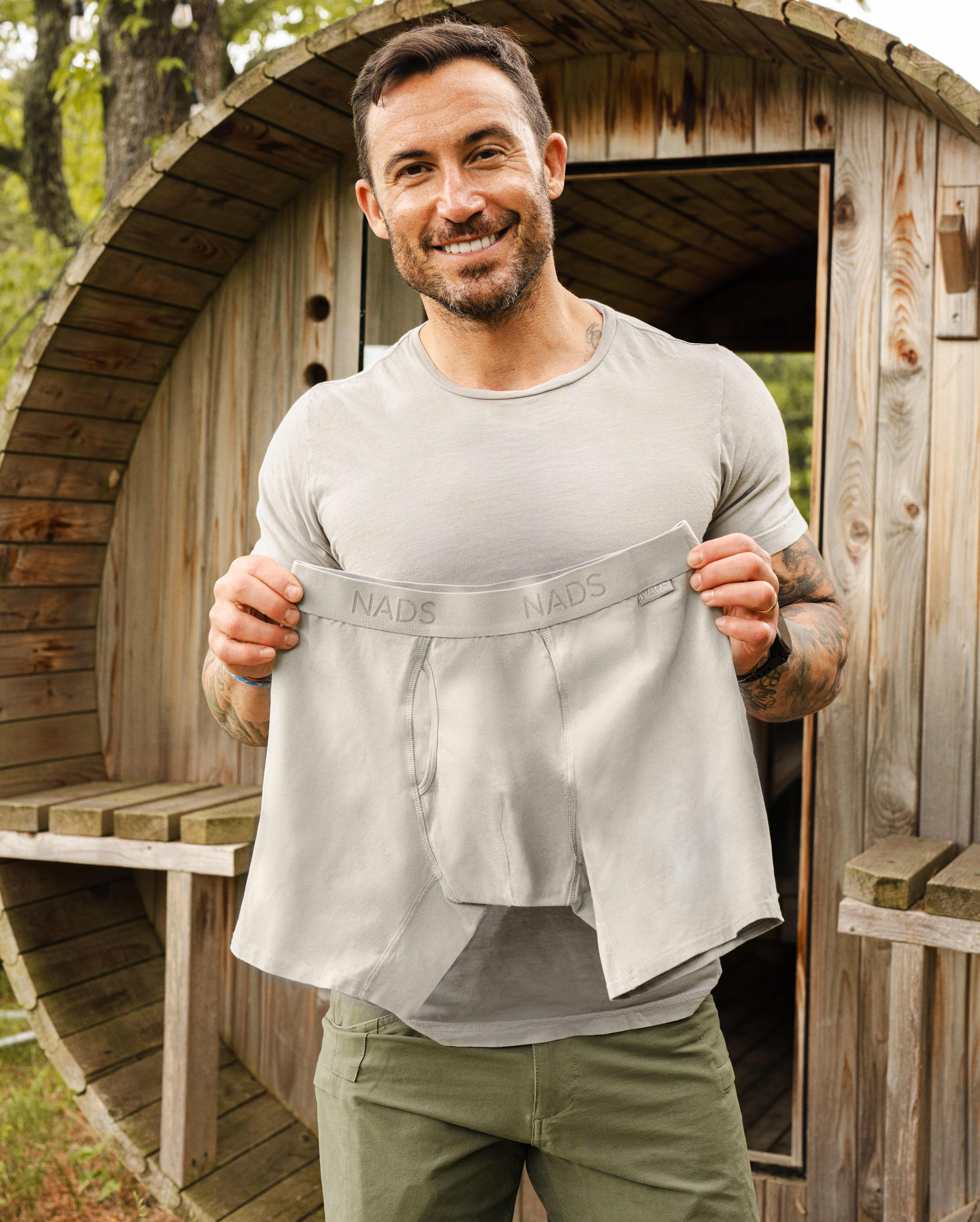 Man holding a pair of gray shorts in front of a wooden structure.
