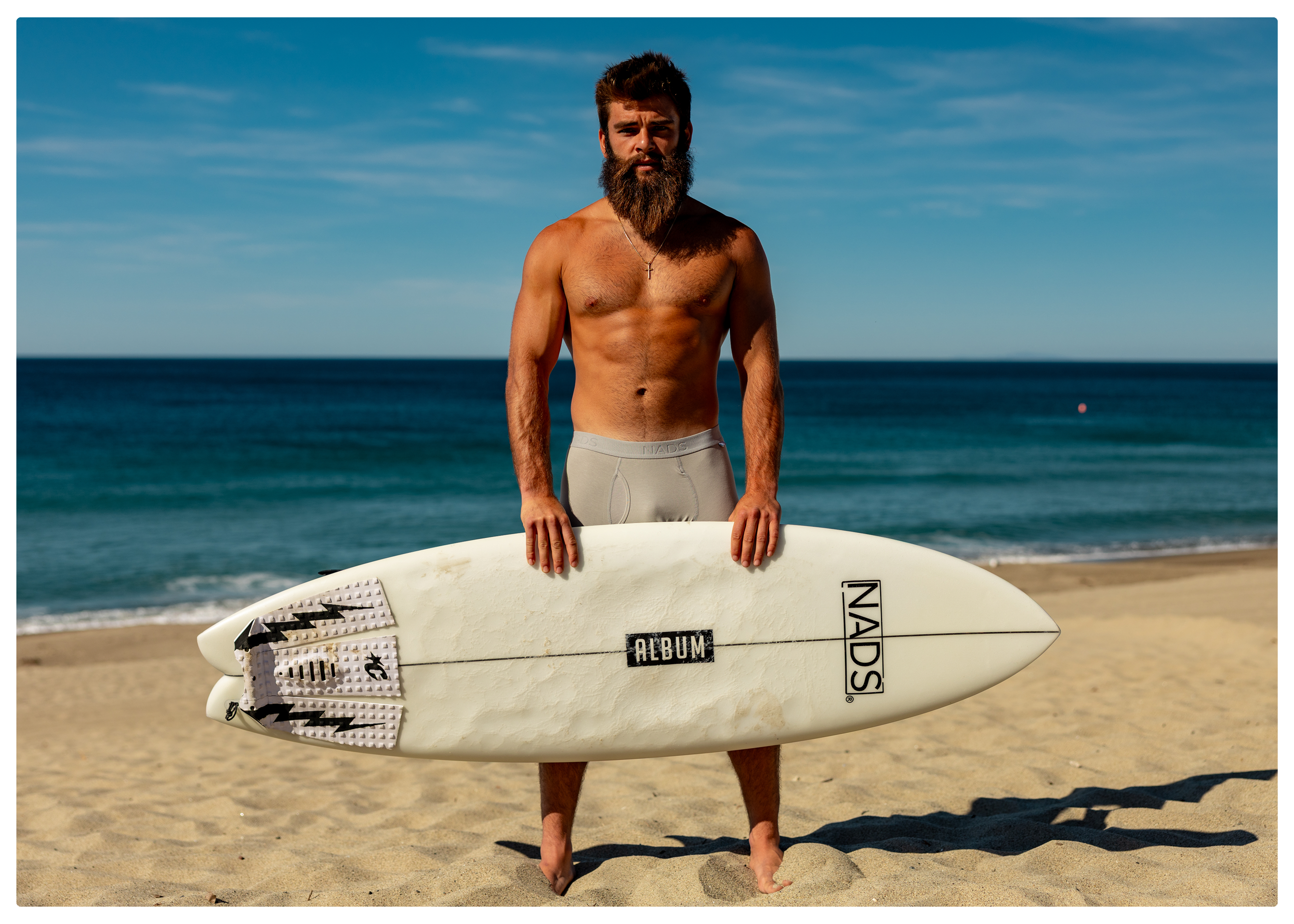 A shirtless man with a long beard stands on a beach holding a surfboard in front of him.