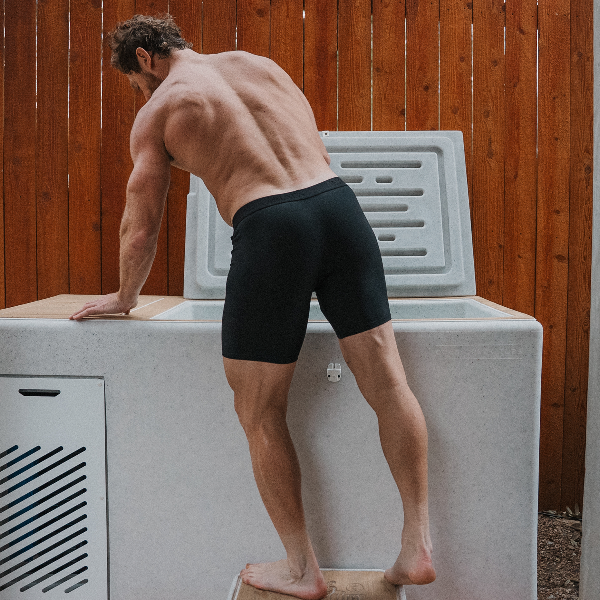 A muscular man, seen from the back, leans over a large, light-colored tub outdoors against a wood fence.
