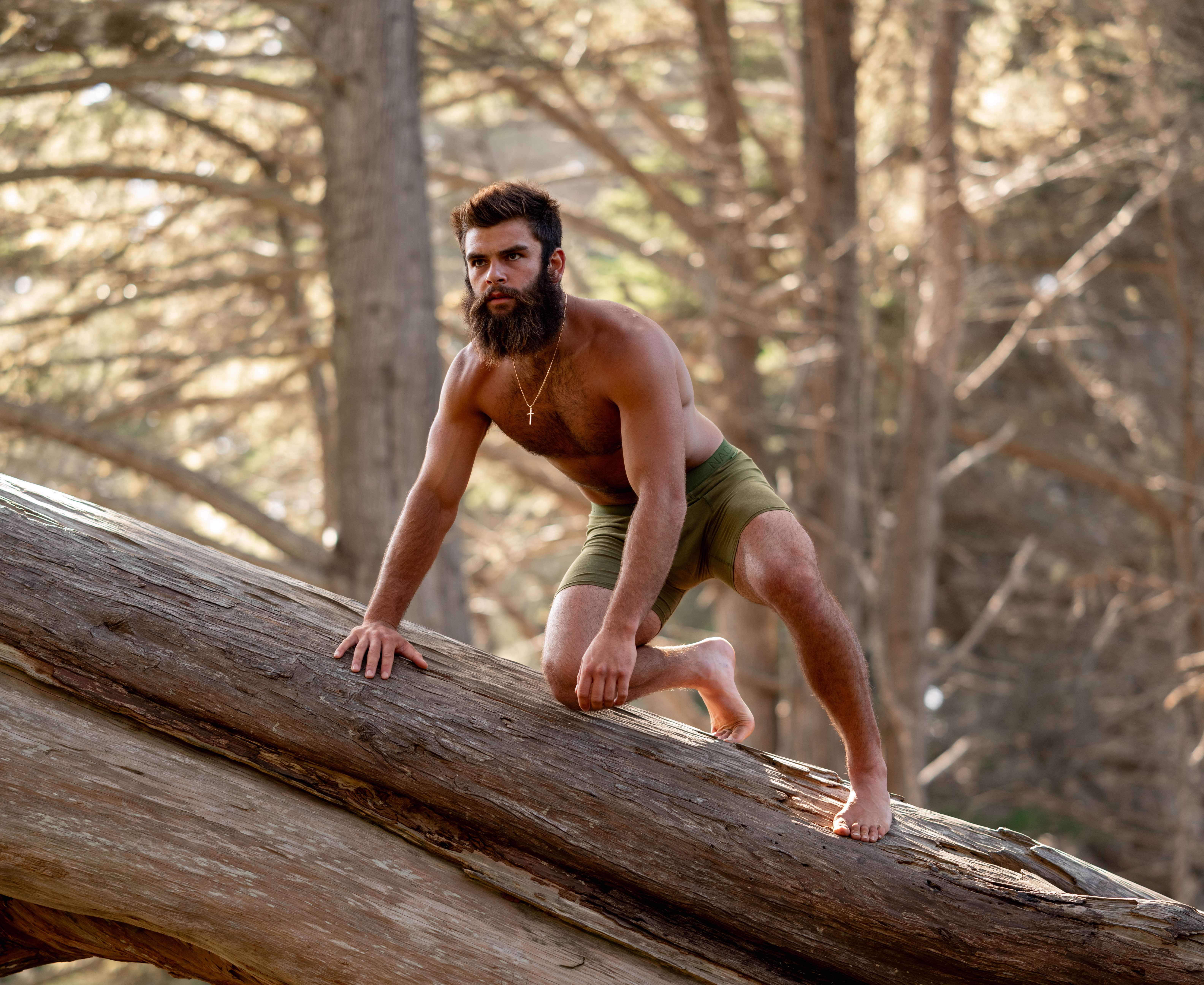 Man with a beard climbing a large tree trunk in a forest.