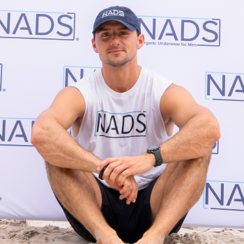 A man in a NADS branded hat and sleeveless shirt sits on the sand in front of a backdrop.