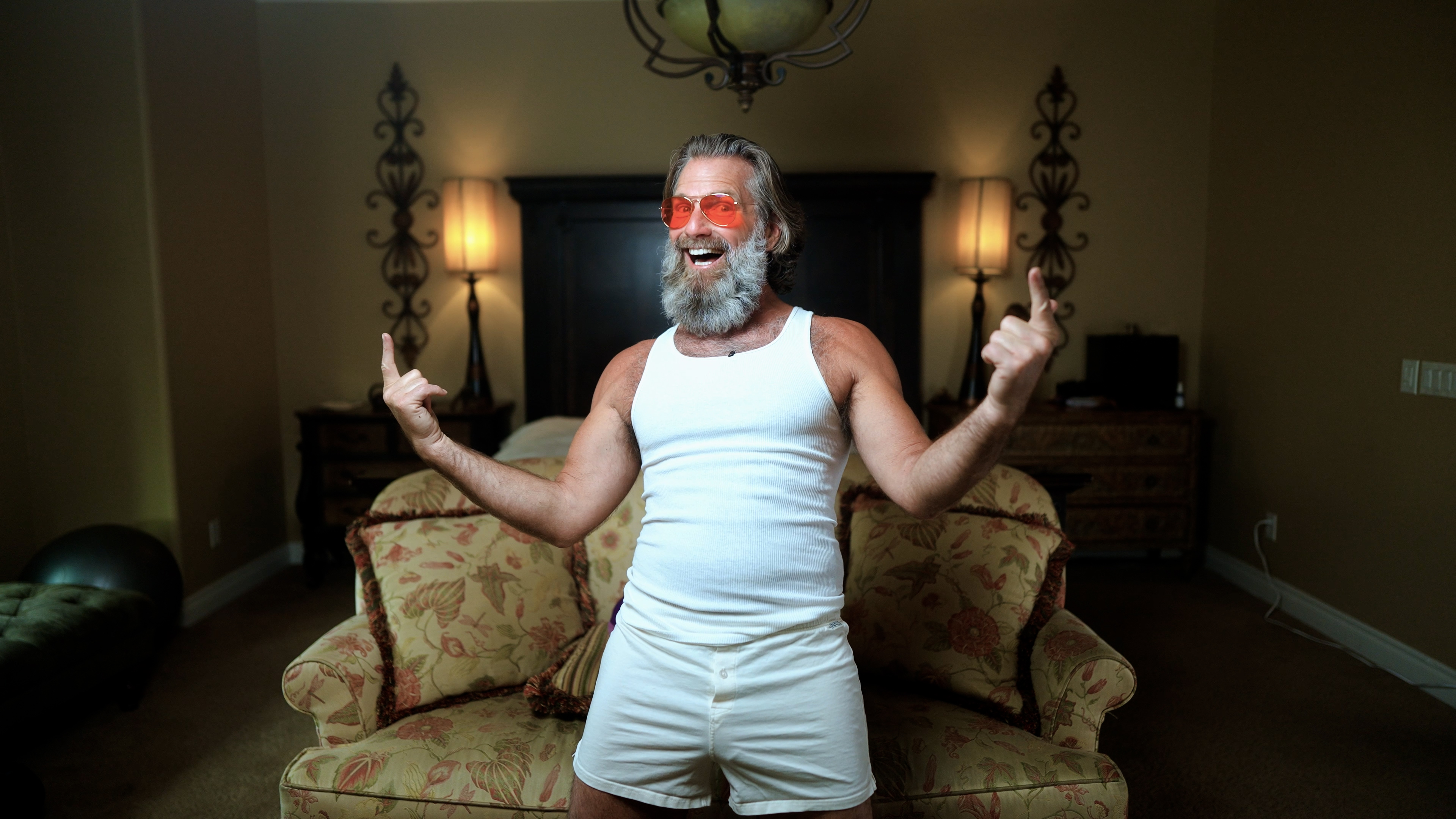 Man with beard and sunglasses smiling, wearing white tank top and shorts, in a living room.