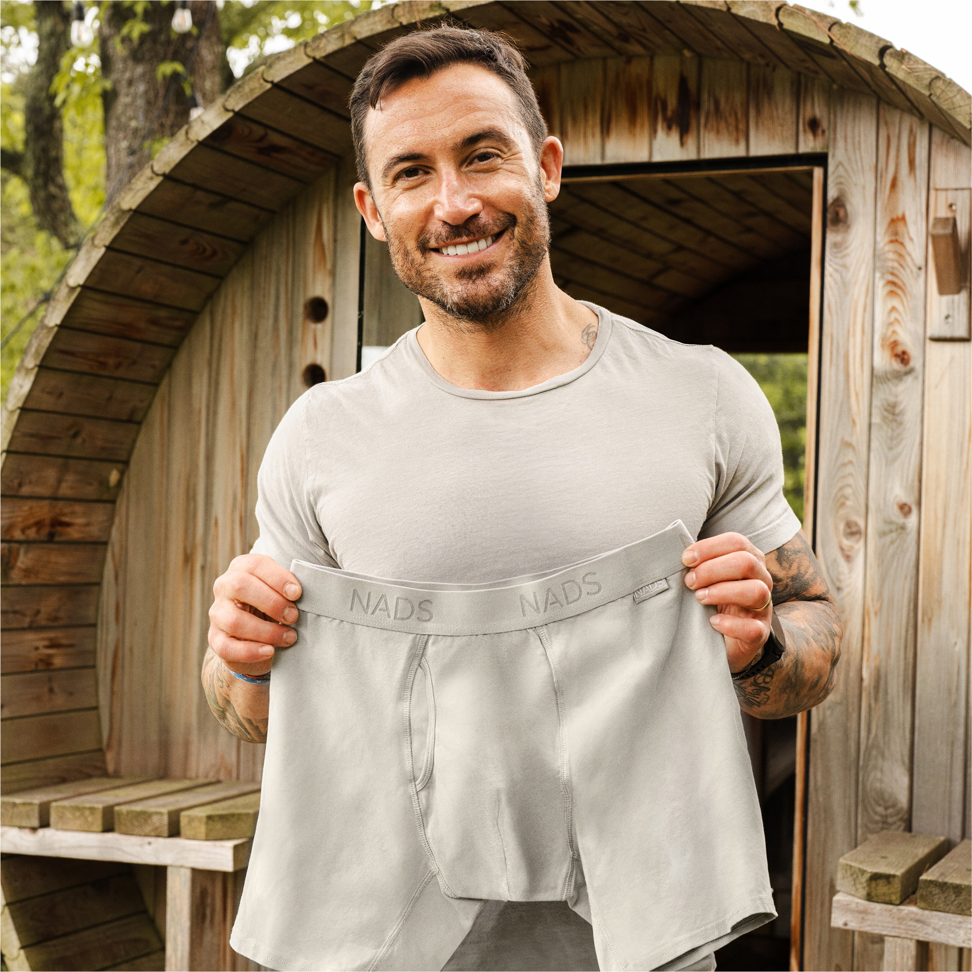 Man smiling, holding a pair of gray shorts in front of a wooden structure.