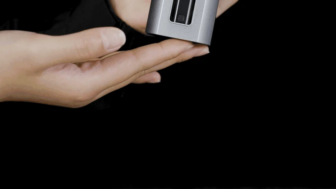 Hand holding a silver rectangular device with a camera lens, black background.