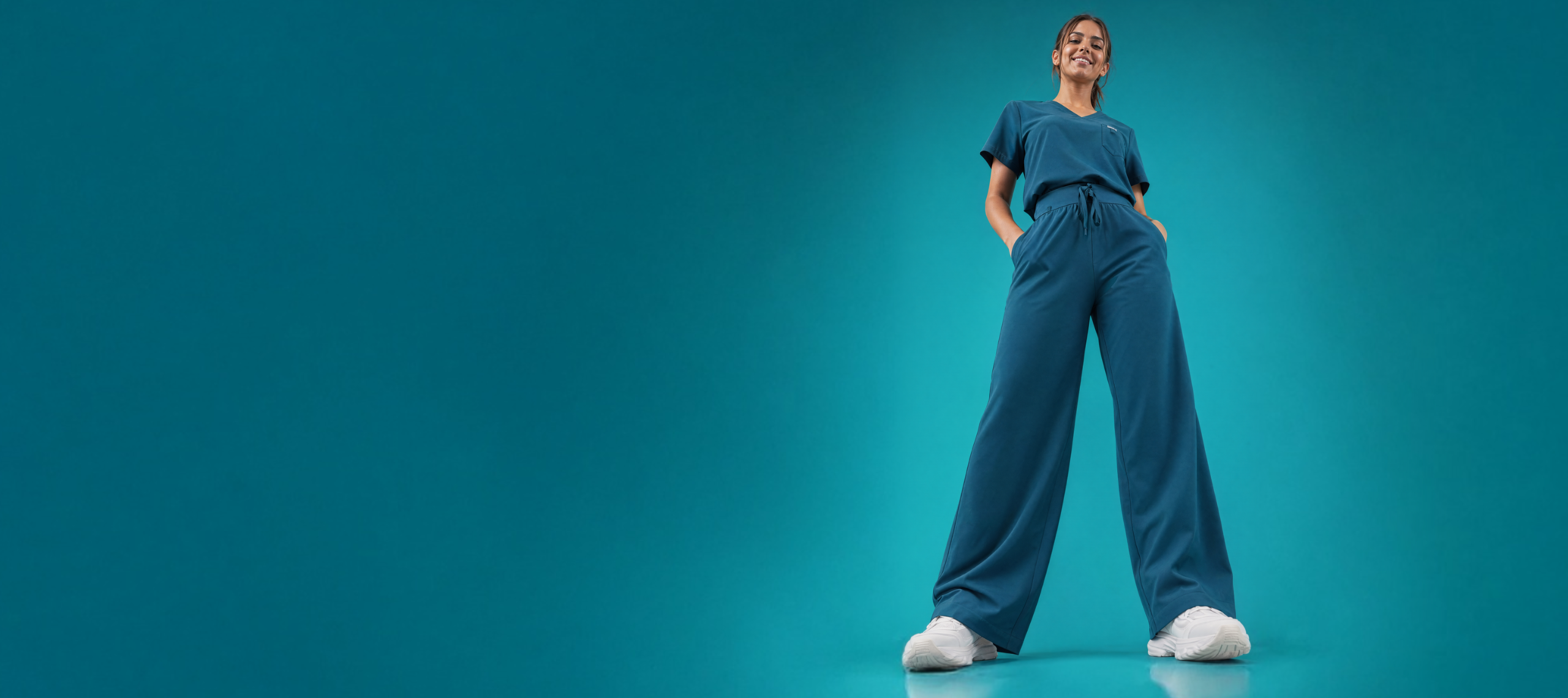 A woman in teal scrubs and white sneakers smiles, posing against a solid teal background.