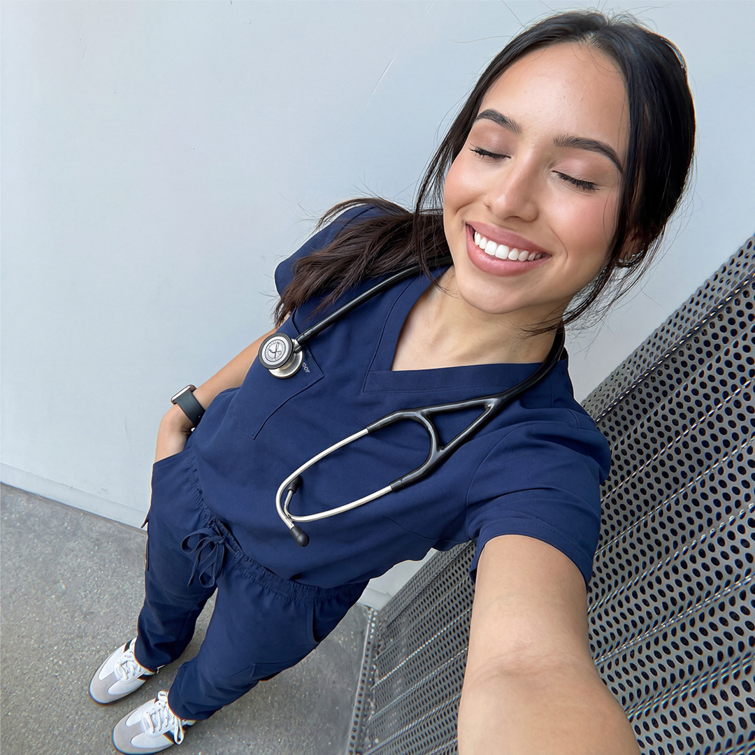 A high-angle selfie of a smiling woman in blue scrubs with a stethoscope around her neck.