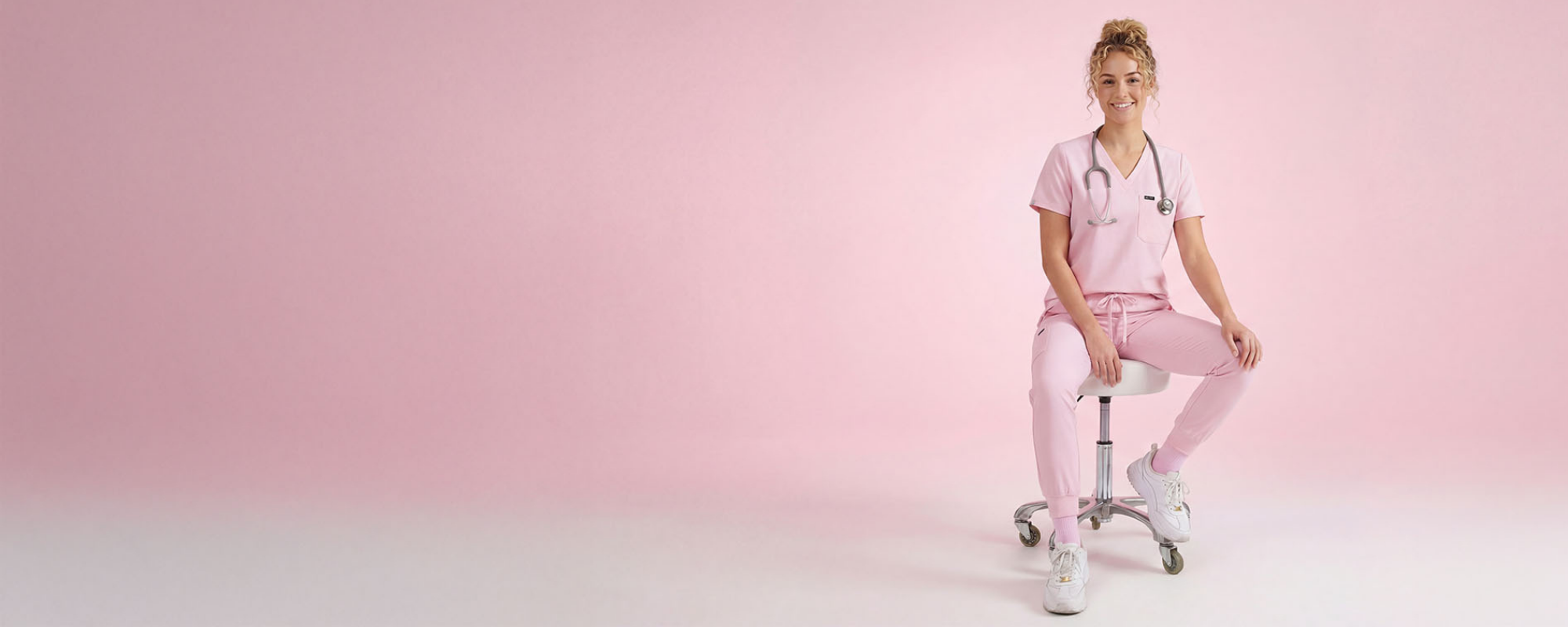 A smiling healthcare professional wearing light pink medical scrubs and white sneakers sits on a rolling stool against a soft pink studio background, with a stethoscope around her neck in a clean, minimalist setting.