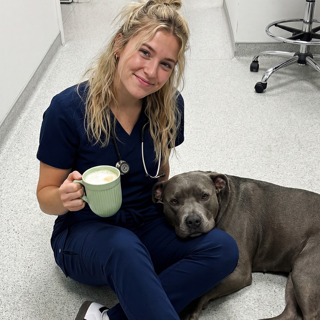 A woman in scrubs sits on a floor holding a mug with a grey dog resting on her lap.